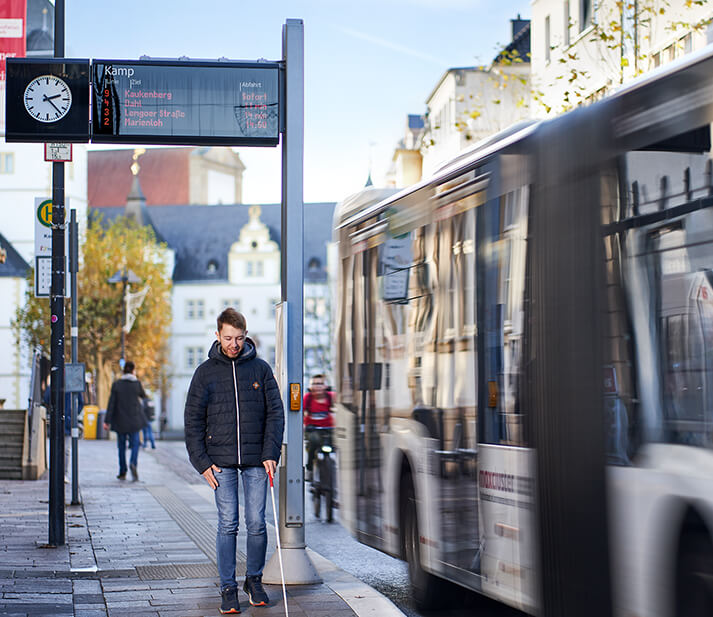 Das Bild zeigt eine sehbeeinträchtigte Person an einer Bushaltestelle mit Fahrgastanzeige.